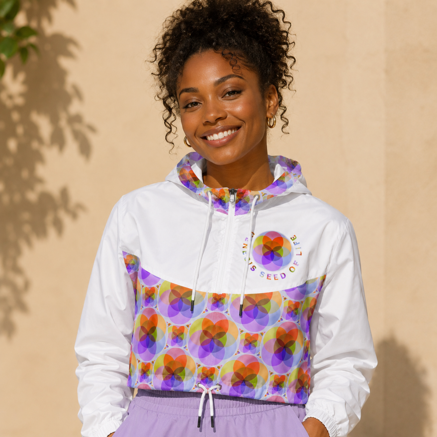 A smiling woman with curly hair wears a white, purple, and orange geometric pattern windbreaker jacket and light purple pants against a beige wall.
