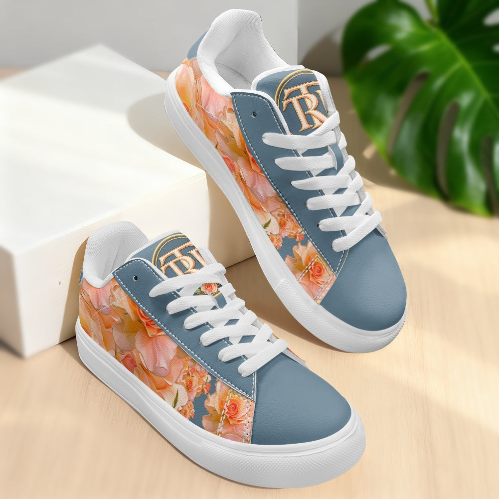 A pair of low-top sneakers with a dusty blue base and a vibrant orange and pink floral print on the sides, white laces, and a gold monogram logo on the tongue. They are displayed on a light wood surface next to a white block and a green plant leaf.