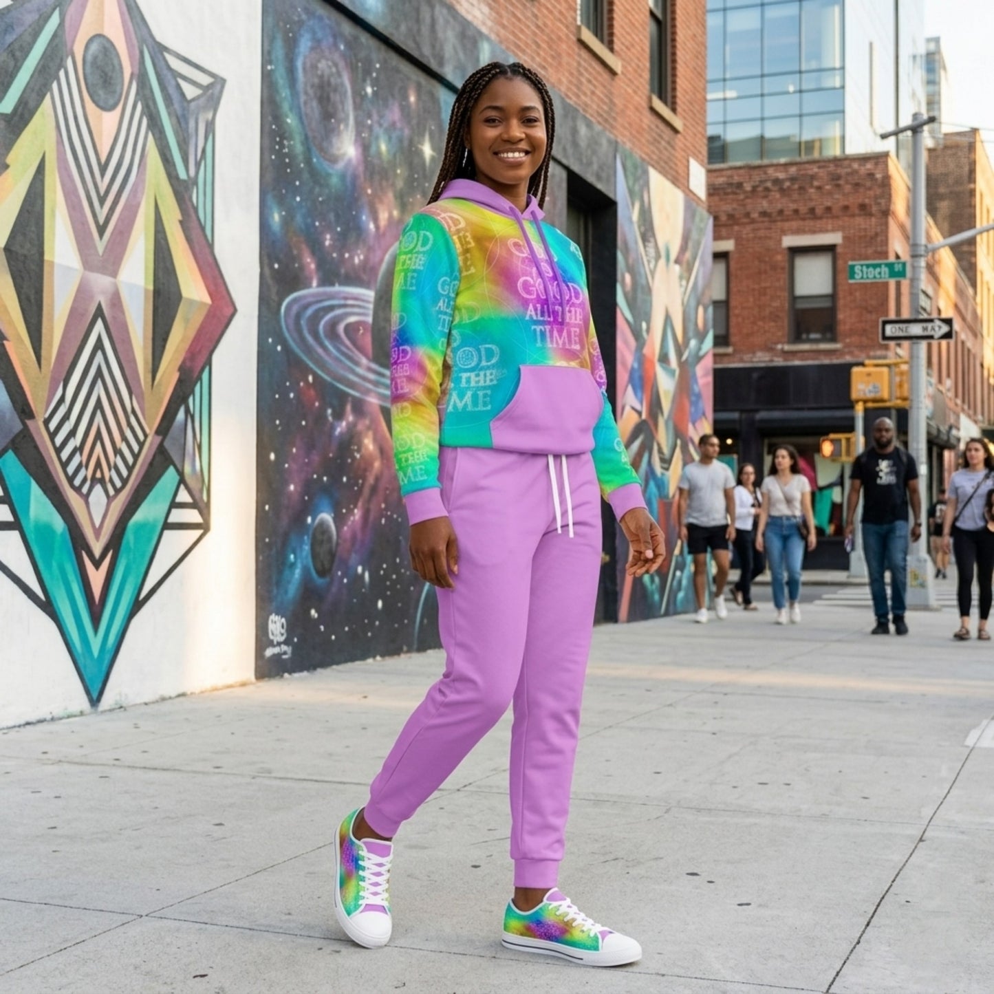 A smiling woman posing on a city sidewalk wearing a purple tracksuit and a tie-dye hoodie with "God is Good All the Time" across the front.