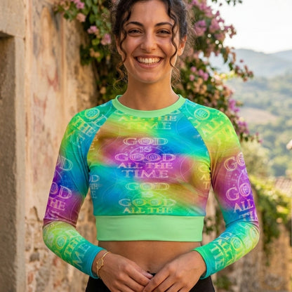 Front view of a woman in a lime green tie-dye crop top with "God is Good All the Time" text, standing against a rustic stone wall with pink flowers.
