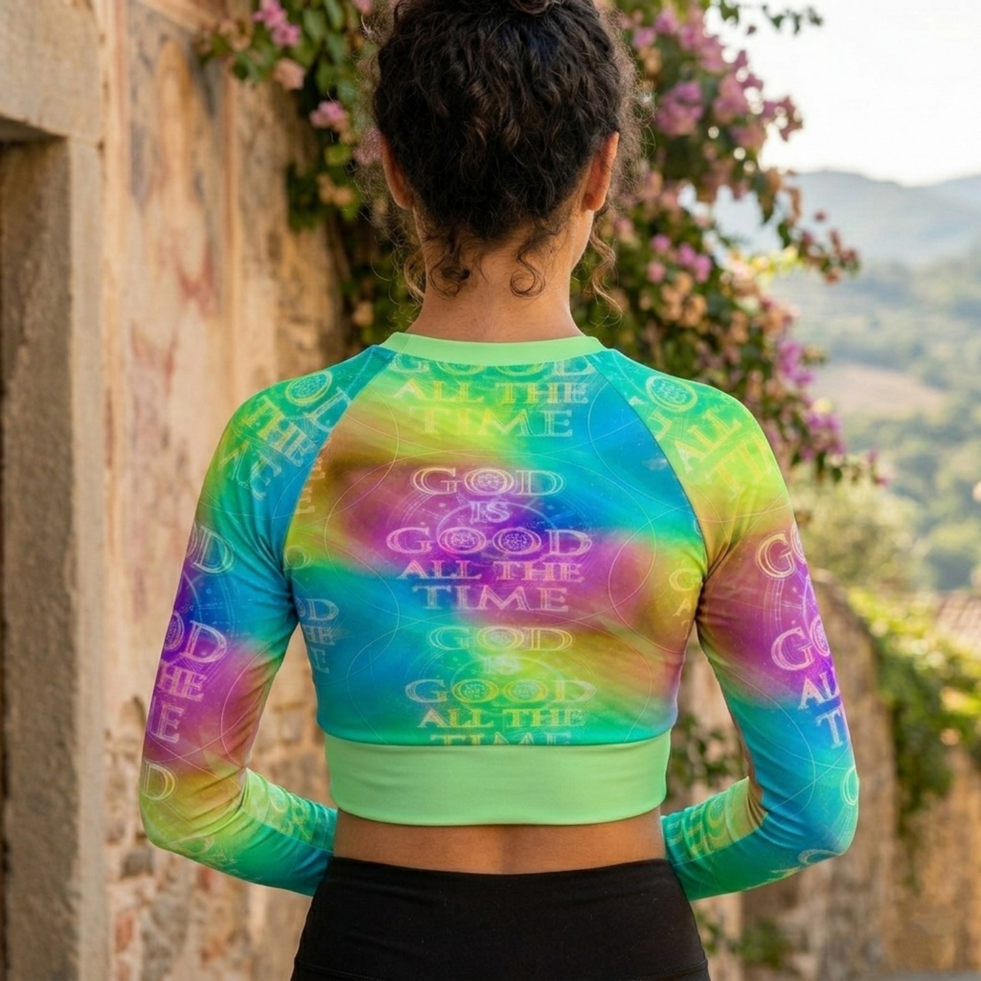 Back view of a woman in a lime green tie-dye crop top with "God is Good All the Time" text, standing against a rustic stone wall with pink flowers.