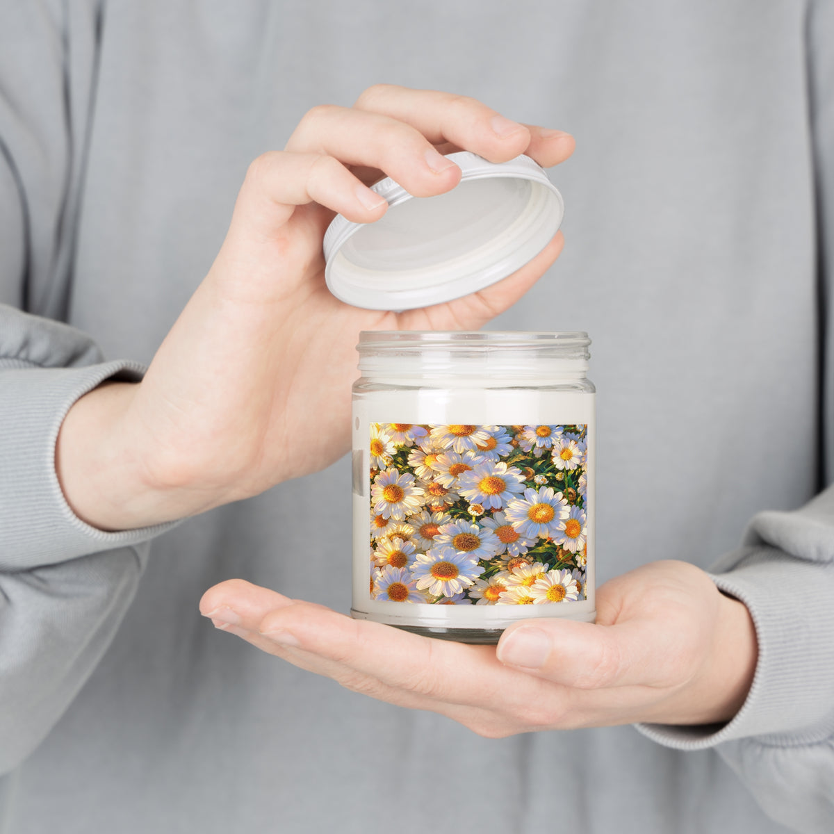 Person holding an opened The Dawning Field 9oz scented soy candle in a clear glass jar with the white lid lifted.