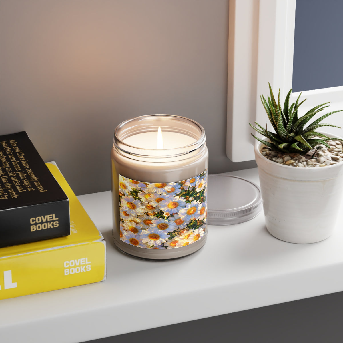Lit The Dawning Field 9oz soy candle jar displayed on a white shelf next to yellow books and a small potted succulent.