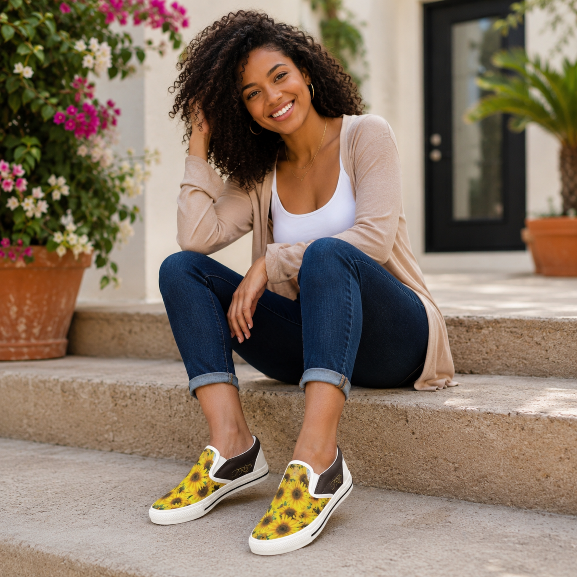 A smiling woman sitting on outdoor steps is wearing TRP Sunflowers in Symmetry women's slip-on canvas shoes and a casual cream cardigan outfit.