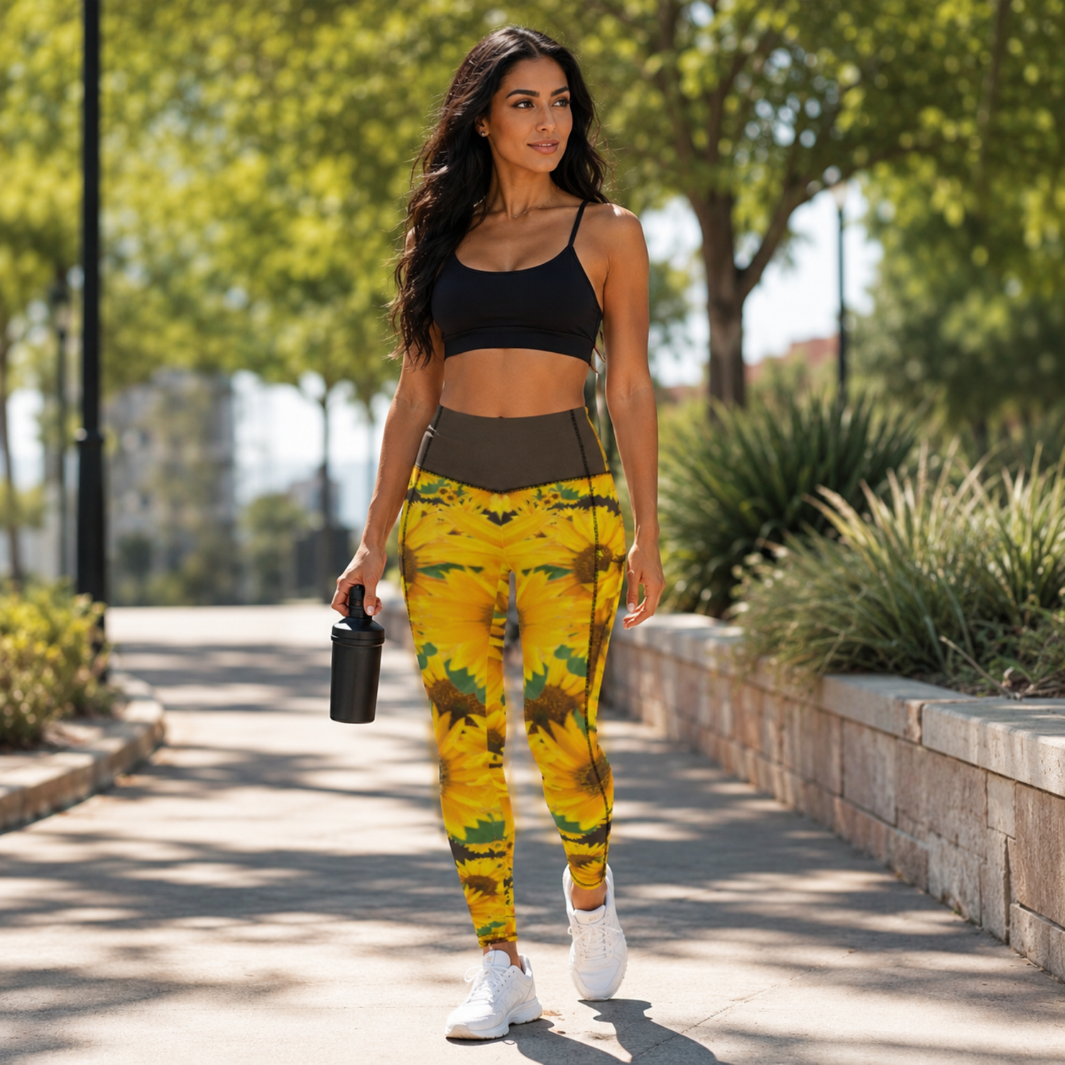 A woman walks forward on a park path wearing TRP sunflower print pocket leggings with a bistre/dark brown waistband, a black sports bra, and white sneakers.