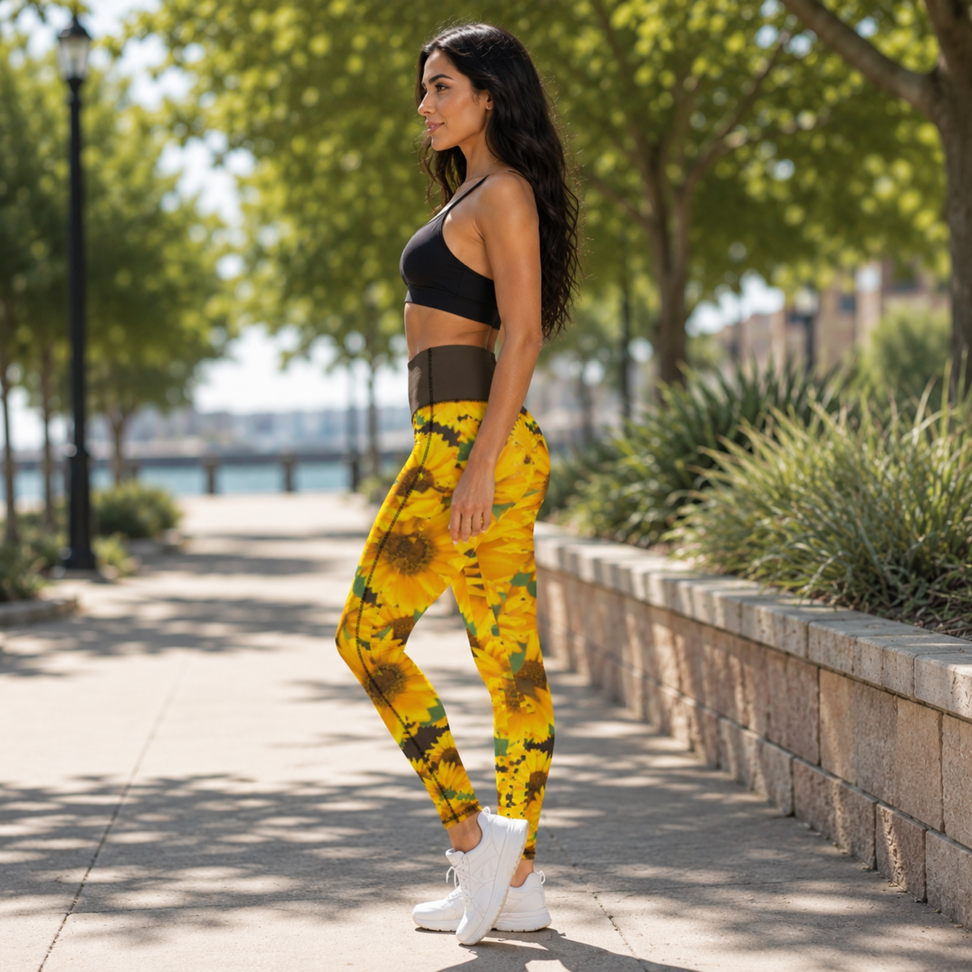 A left-side view of a woman standing on a park pathway wearing symmetrical sunflower-print high-rise pocket leggings with a bistre/brown waistband and black sports bra.