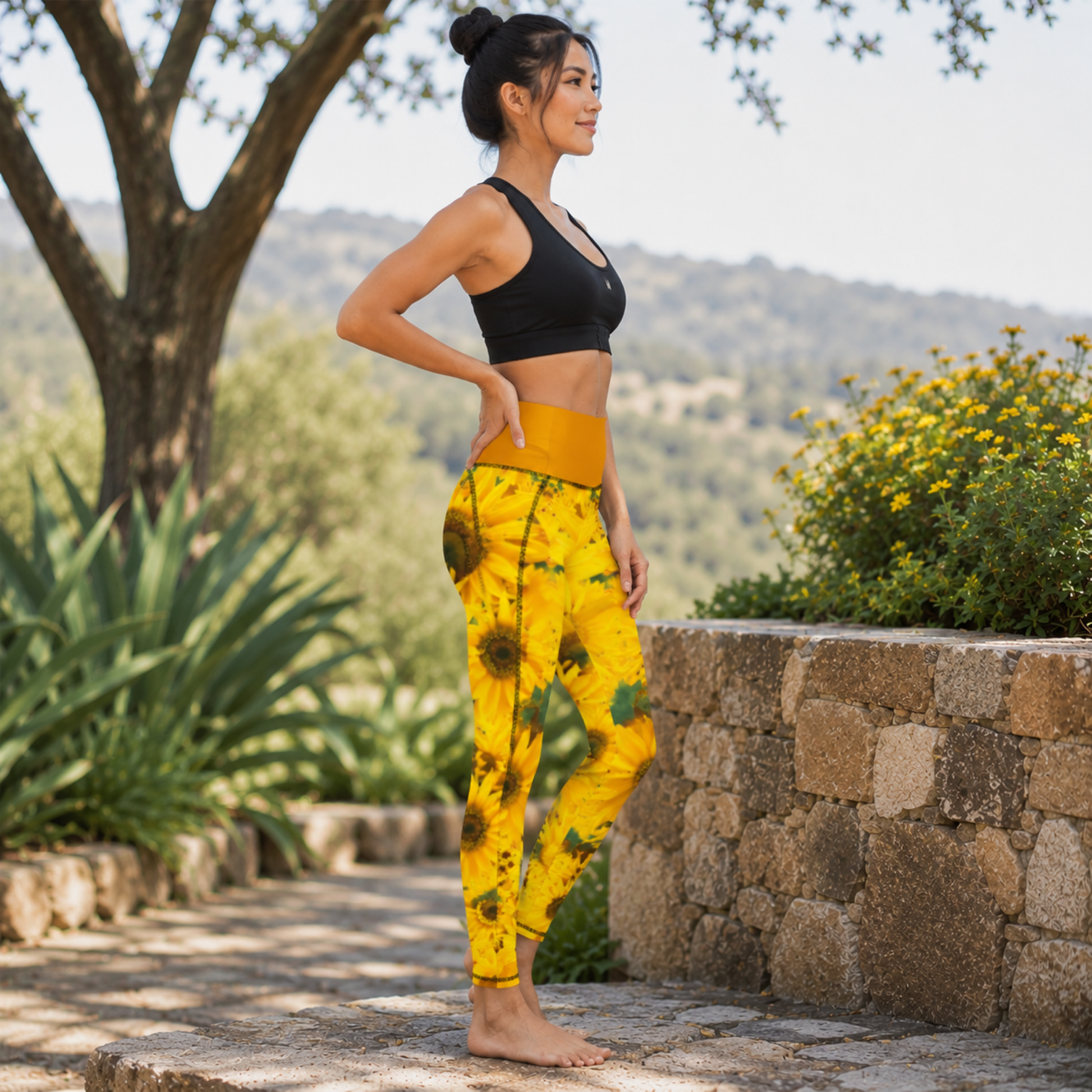 Right-side view of a woman standing barefoot near a stone wall, wearing sunflower-print pocket leggings with a dark tangerine/mustard yellow waistband and a black sports bra.