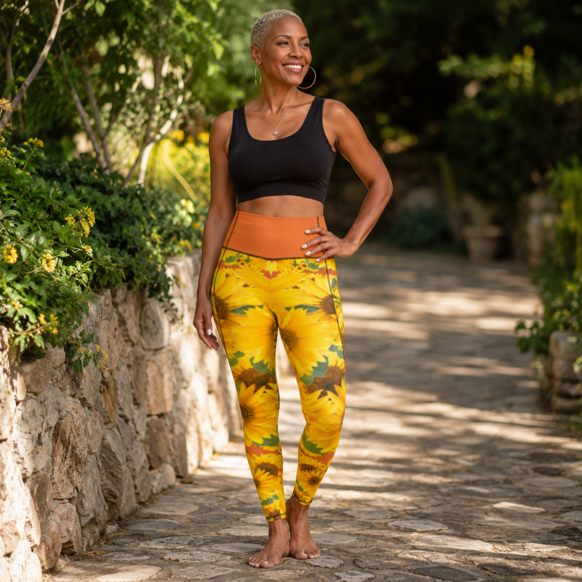 Front view of a Black woman with short blonde hair posing barefoot in sunflower print pocket leggings featuring a bright vivid red tangelo/orange waistband.